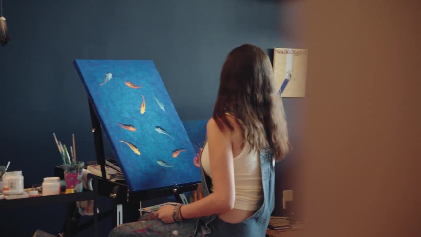 A talented young artist sits in her creative studio, painting a vibrant canvas of colorful fish swimming in deep blue water