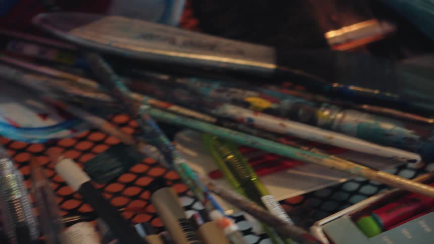 A close-up slow-motion shot pans across various used paintbrushes and other art supplies on a cluttered work table