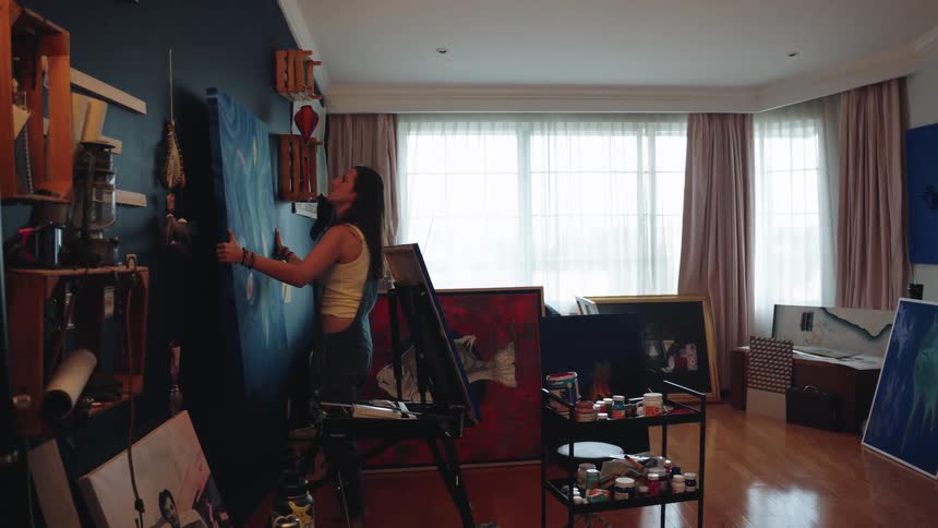 A young female artist carefully lifts a large canvas and places it on the wall of her messy home studio