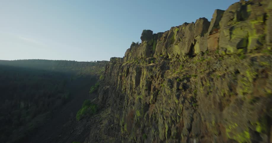 A dramatic aerial view follows a mossy, jagged cliff face rising from a forested valley during a spectacular sunrise