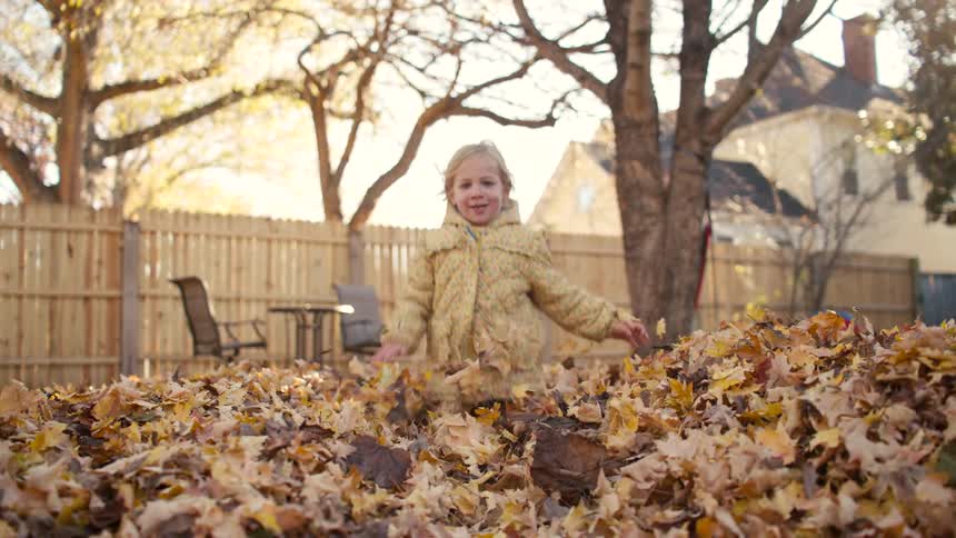 A happy little girl in a yellow jacket joyfully runs through a pile of fallen autumn leaves toward the camera in slow motion