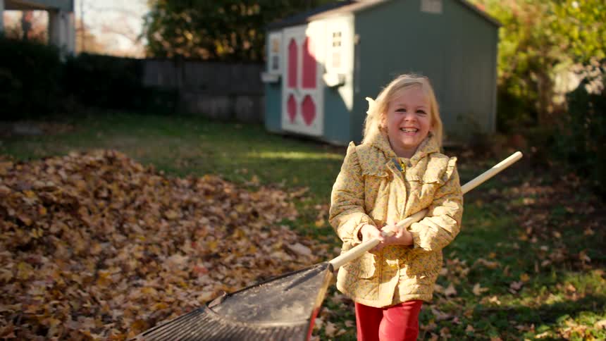 A joyful young girl laughs as she runs toward the camera in slow motion, playfully raking colorful autumn leaves in a sunny backyard