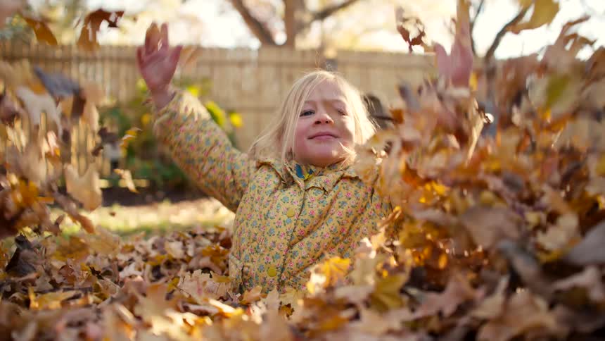 A happy little girl smiles as she sits in a pile of autumn leaves and throws them up into the air in slow motion