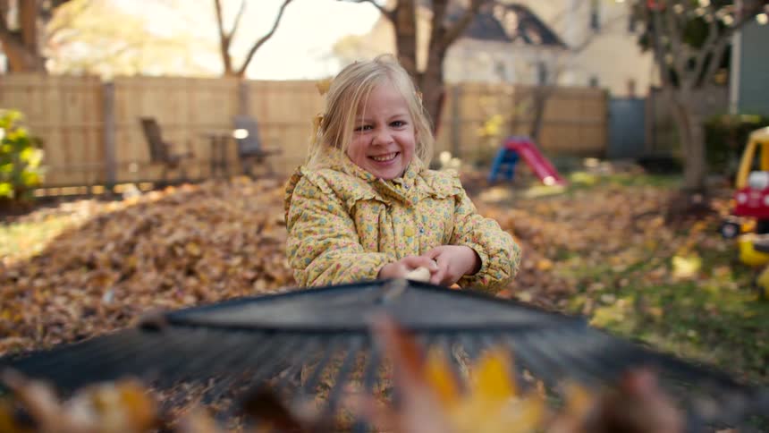 A joyful little girl with a big, happy grin energetically rakes autumn leaves toward the camera in her sunny backyard