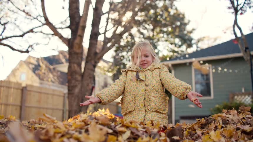 A joyous young girl throws a pile of autumn leaves into the air in slow motion in a sunny backyard