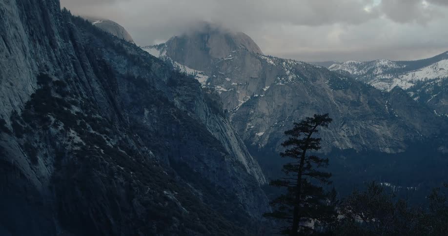 A majestic, moody view of Yosemite National Park, slowly tilting up to reveal Half Dome and the surrounding snow-dusted mountains under a cloudy sky