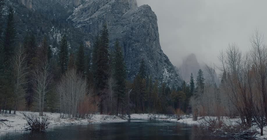 A calm river reflects a snow-dusted forest before tilting up to reveal a towering, misty mountain peak in winter