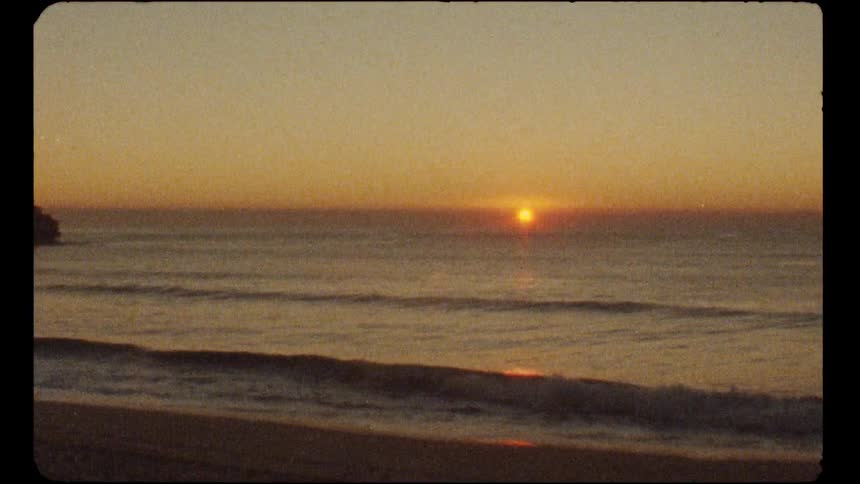 Nostalgic 8mm film captures a serene sunrise over the ocean as waves gently roll onto the shore