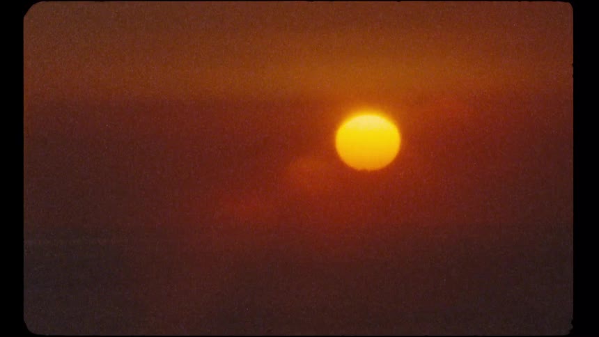 A grainy, vintage film close-up captures the glowing yellow sun in a hazy orange sky at sunset