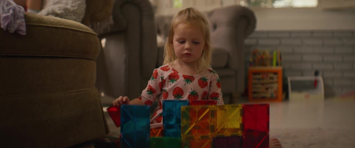 A cute little blonde girl in strawberry pajamas builds with colorful magnetic tiles on the floor