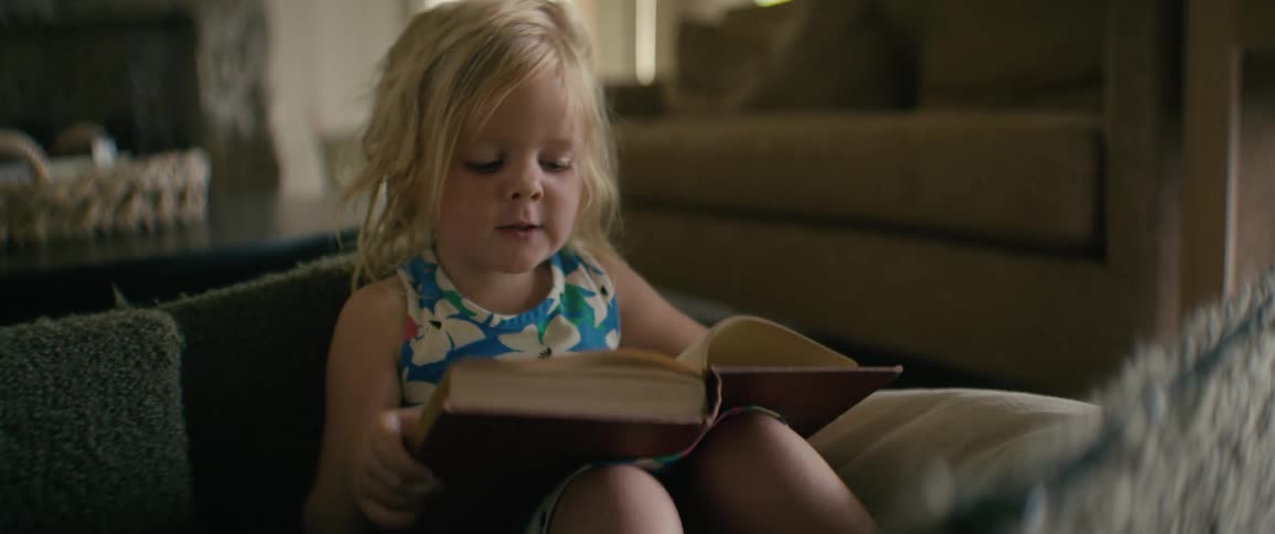 A cute blonde toddler sits in her living room, engrossed as she pretends to read a big storybook
