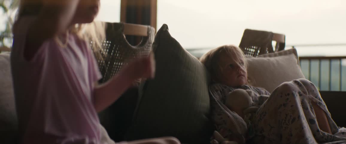 A young girl holds her doll while resting on a cozy couch as her sister attentively watches something offscreen