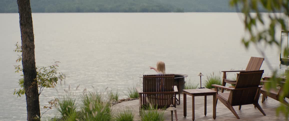 A young blonde girl sits on a quiet lakeside patio, pointing out at the calm water