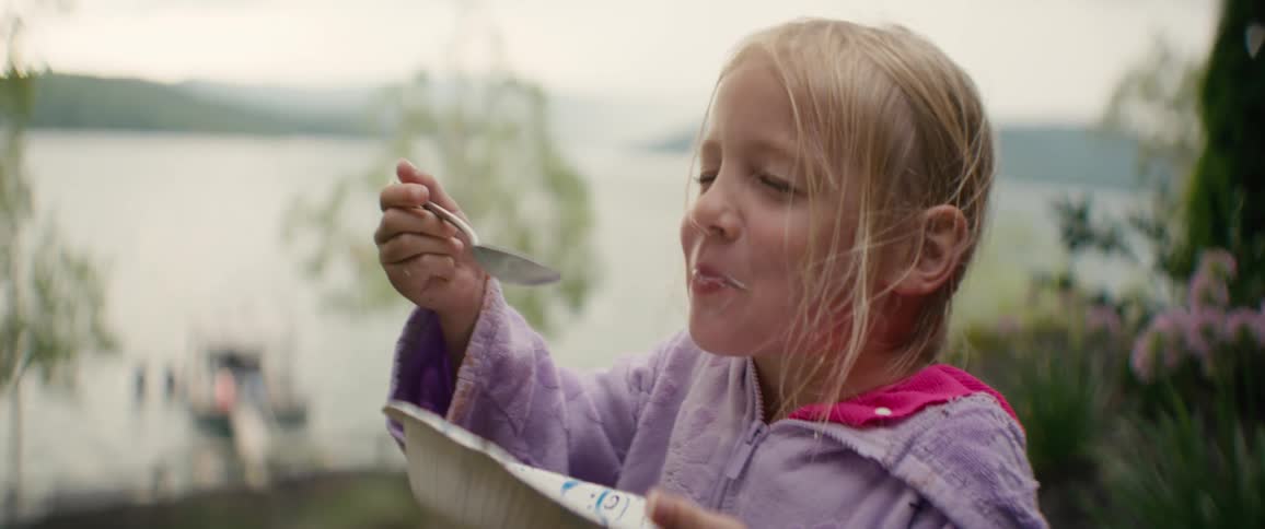 A cute blonde girl in a purple robe happily eats from a bowl, smiling with a yogurt mustache with a lake in the background