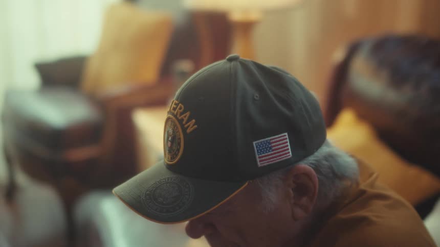 A pensive elderly American veteran wearing a hat with an American flag sits alone on a couch