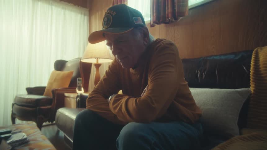 A lonely elderly veteran watches a tablet on his couch with a pensive expression in a dimly lit living room