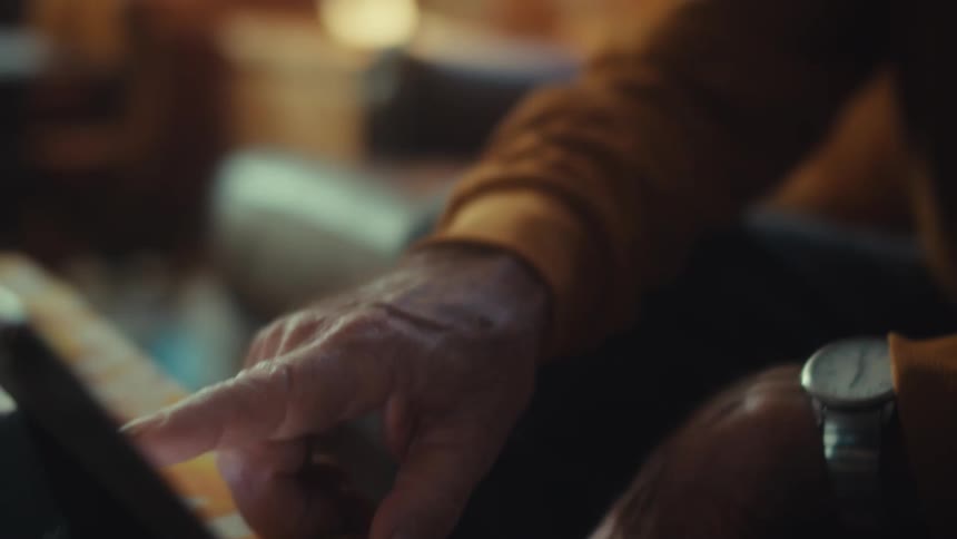 A close-up shot reveals an older man's wrinkled hand swiping on a tablet before the camera slowly tilts up to his focused face