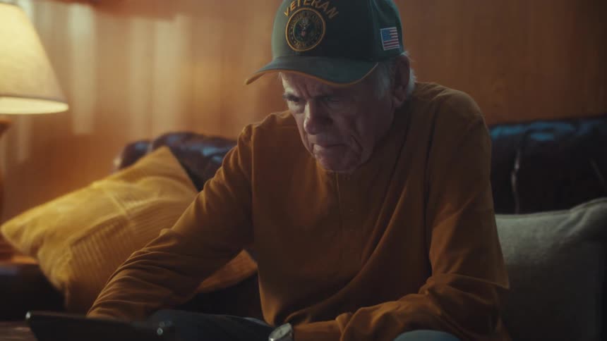 A slow zoom reveals an elderly veteran with a focused and emotional expression as he uses a tablet on his living room couch