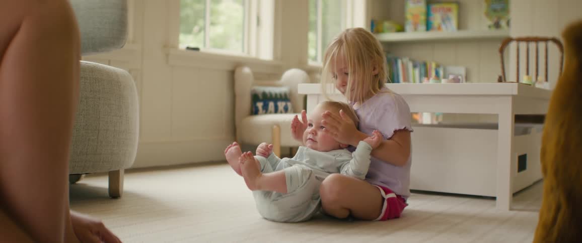 A loving older sister plays a gentle game of peekaboo with her happy baby sibling on the floor of their bright living room