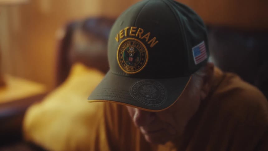 An elderly US Army veteran wearing a patriotic hat looks down with a somber expression in a dimly lit room