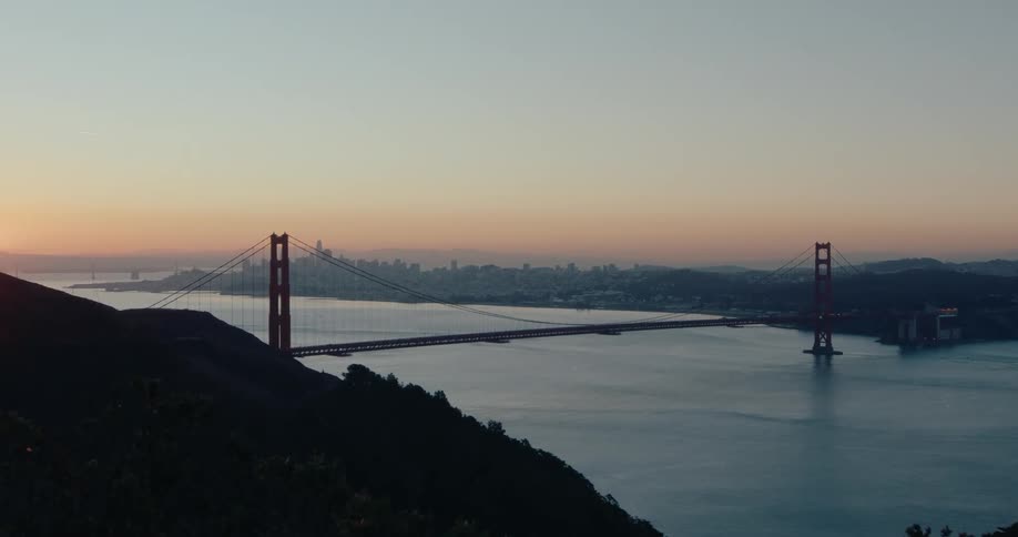 A serene wide shot captures the iconic Golden Gate Bridge and the distant San Francisco skyline as the sun rises over the calm bay waters