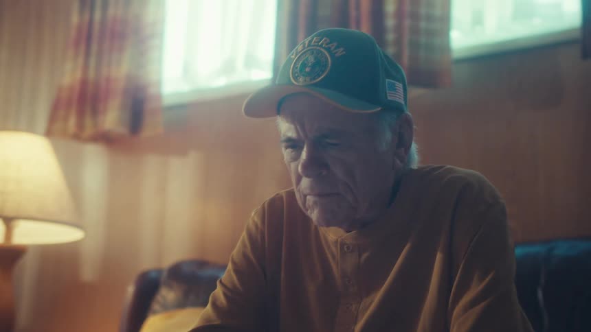 A pensive elderly veteran wearing a cap uses a digital tablet in his dimly lit, cozy living room