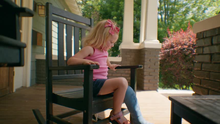 A young girl with a broken leg in a cast carefully gets up from a rocking chair on a sunny porch