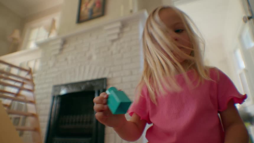 A young girl with blonde hair in a pink shirt concentrates while playing with colorful building blocks in a sunlit living room