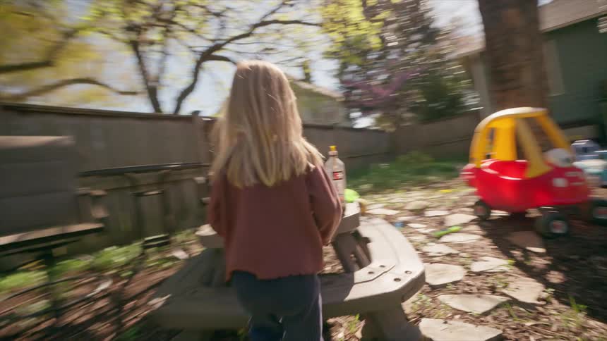 A cinematic slow-motion shot follows a young blonde girl as she runs through a sunny backyard to a picnic table