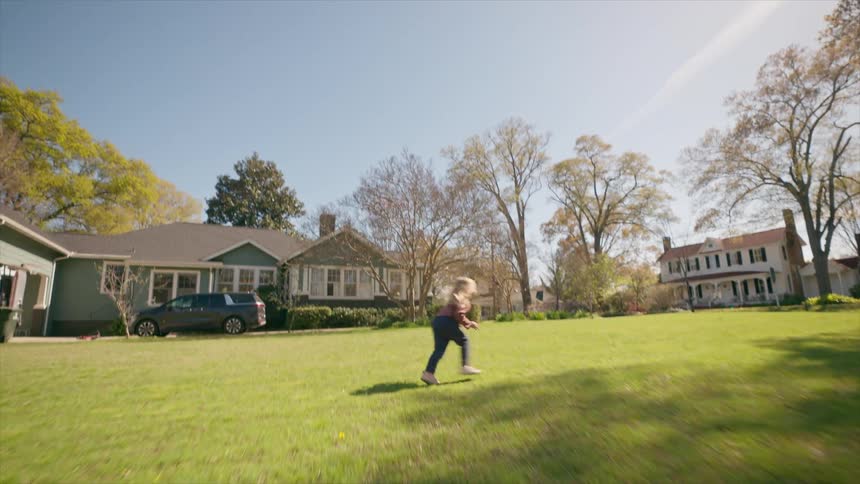 A low-angle shot follows a young girl as she runs across a large grassy yard in a suburban neighborhood, tilting up to the bright blue sky