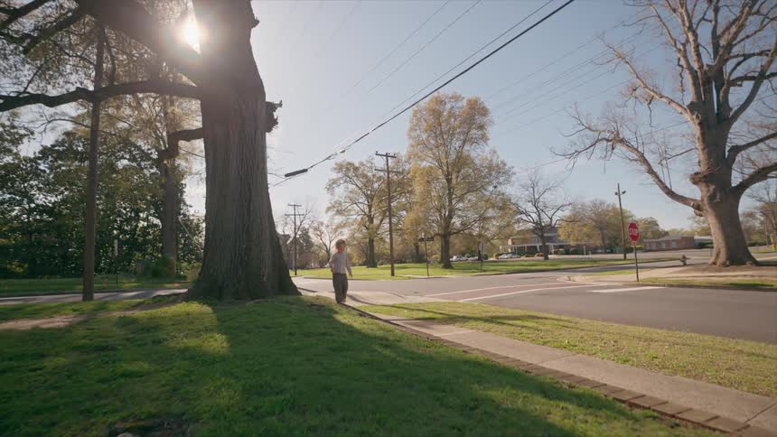 A young child emerges from behind a large tree and walks barefoot down a sunlit sidewalk