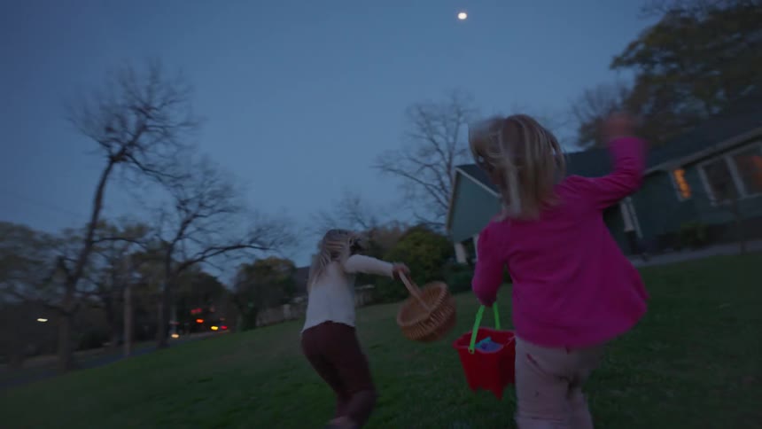 Two joyful young girls run happily with their baskets through a grassy yard at twilight