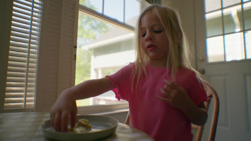 Little girl in a pink shirt enjoying a snack at a sunlit kitchen table