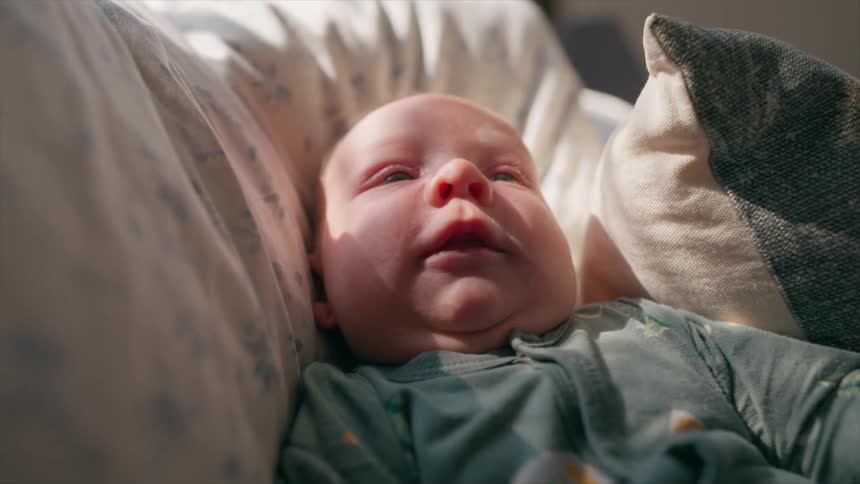 A close-up of a newborn baby crying and then calming down as they lie in a bassinet bathed in warm sunlight