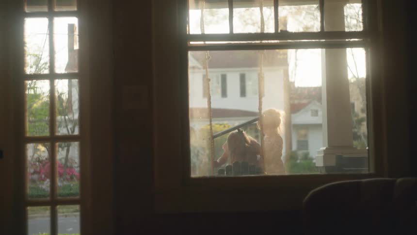 A nostalgic slow-motion view from inside a home looking out at two young children playing on a porch swing during golden hour