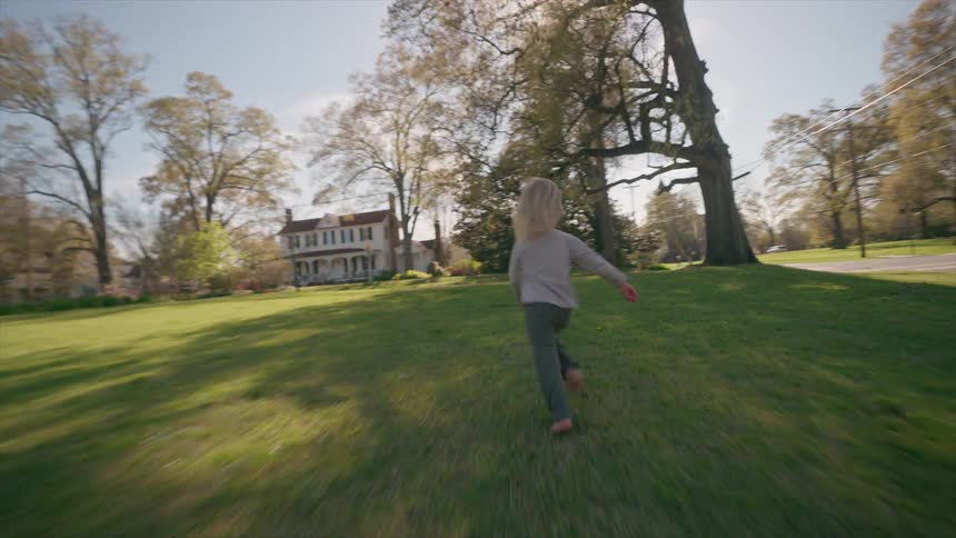 Two young blonde sisters play and run barefoot across a sunny green lawn in their suburban neighborhood