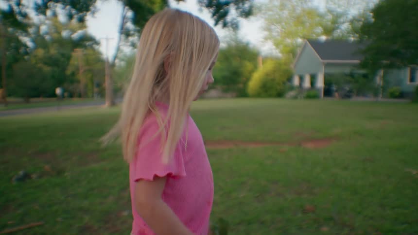 A young blonde girl in a pink shirt walks through a grassy yard while holding a large green leaf
