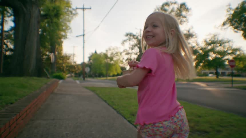 A joyful blonde girl in a pink shirt runs along a neighborhood sidewalk, turning back to smile at the camera during a beautiful sunset