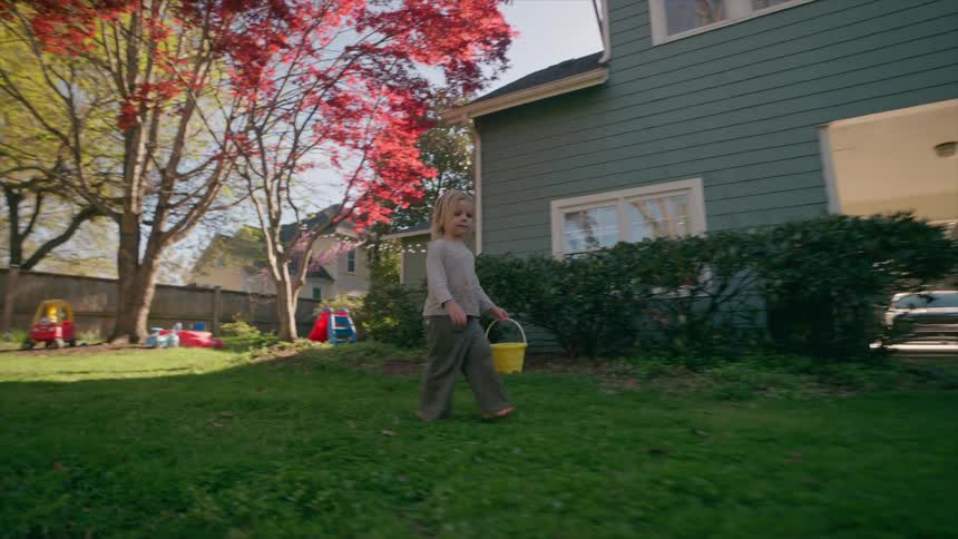 A barefoot toddler girl with a yellow bucket walks across the lush green grass of her backyard on a sunny spring day