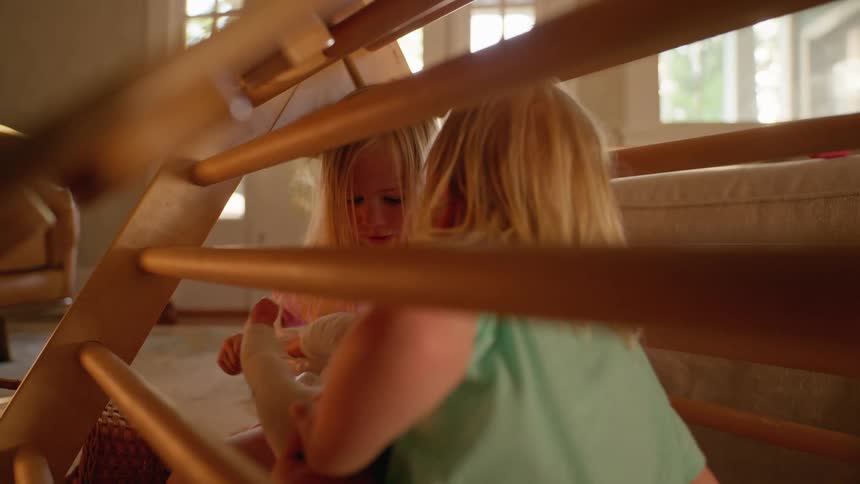Two young sisters laugh and play with dolls under a wooden climbing structure in their sunlit living room