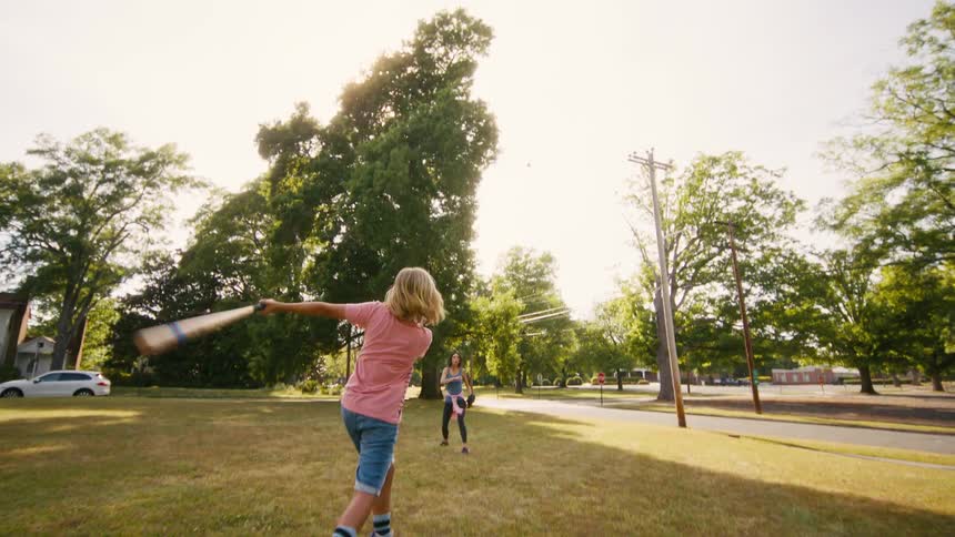 A young boy practices his baseball swing with his mother in a grassy park on a bright, sunny day