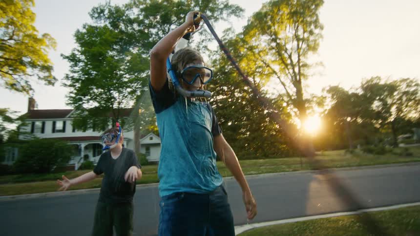 Playful kids in snorkel gear have a water fight with a garden hose in their front yard at sunset