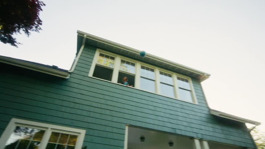 A boy throws a ball up to a girl in a second-story window who then throws it back down in their sunny backyard
