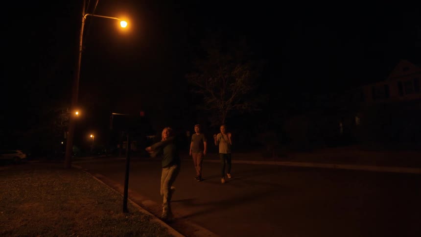A teenage boy violently smashes a mailbox with a baseball bat on a dark suburban street, and then she and her friends run off into the night