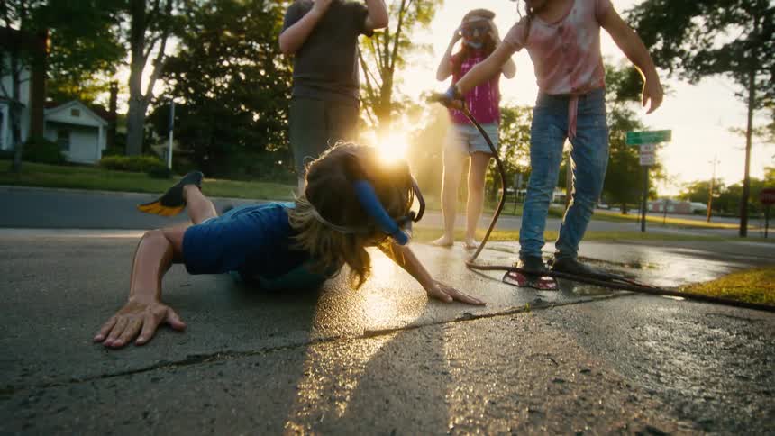 A group of kids wearing snorkel gear have a water fight on their driveway as one pretends to swim on the concrete during a golden sunset