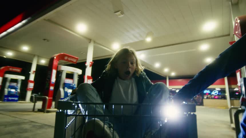 A happy kid gets a thrilling ride in a shopping cart pushed by friends through a gas station at night