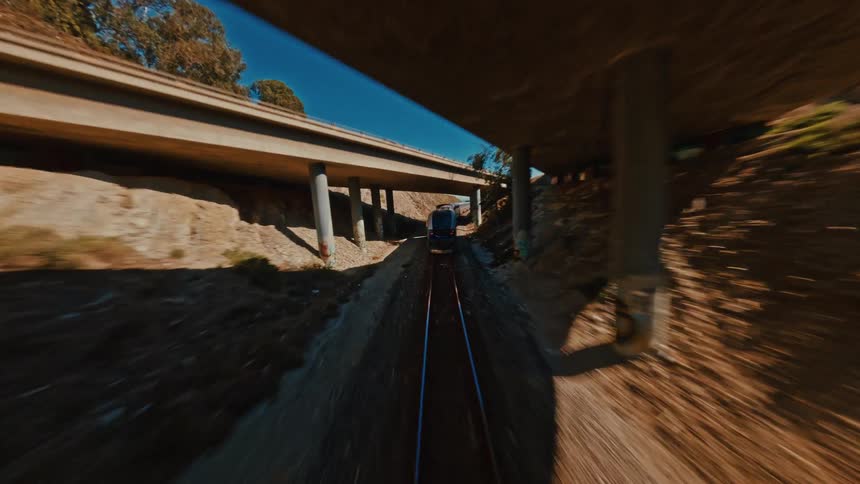 Dynamic FPV drone shot follows a passenger train speeding along tracks and going under a highway bridge