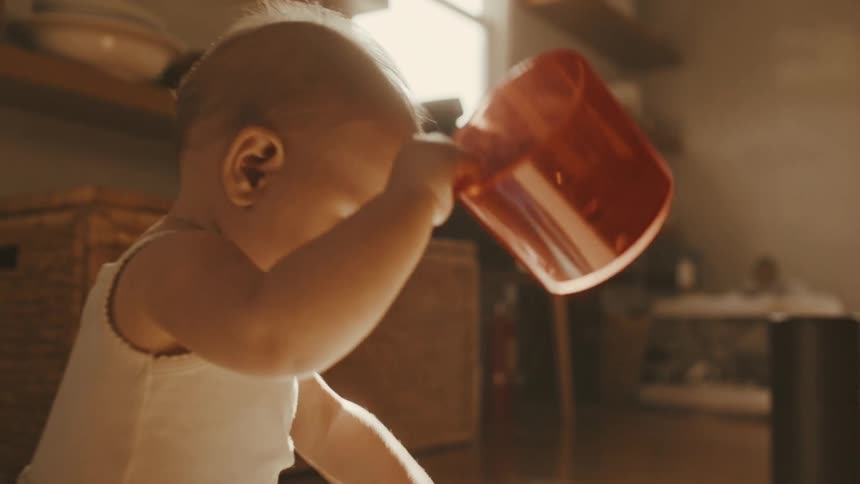 A cute baby in a white tank top sits on the floor in a sunlit room and plays with a red cup in slow motion
