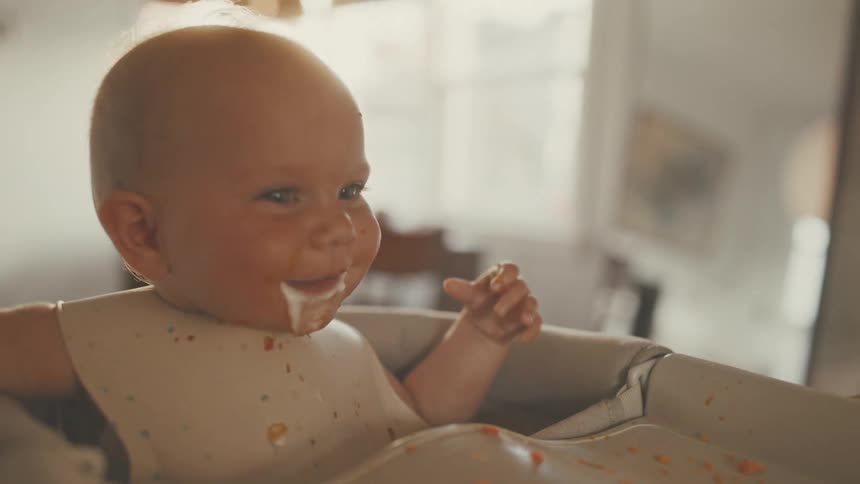A happy, messy baby with food on their face smiles while sitting in a high chair in a sunlit room
