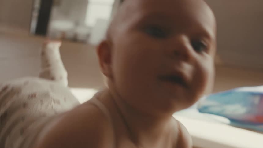 An adorable baby has tummy time on the floor in a sunlit room and looks curiously at the camera in slow motion